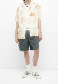 Camicetta beige a maniche corte con bottoni e motivo floreale, abbinata a pantaloni corti verdi. Indossata con calzini bianchi e sneakers crema con dettagli blu.