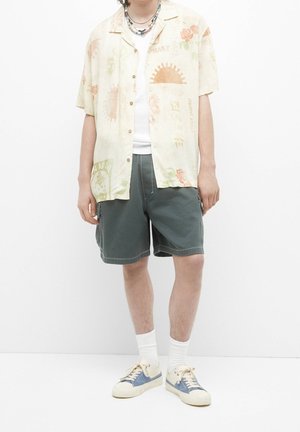 Lyhythihainen, beige nappipaita, jossa on kukkakuosia, yhdistetty vihreisiin shortseihin. Jalassa valkoiset sukat ja vaaleanbeige kengät, joissa on sinisiä yksityiskohtia.