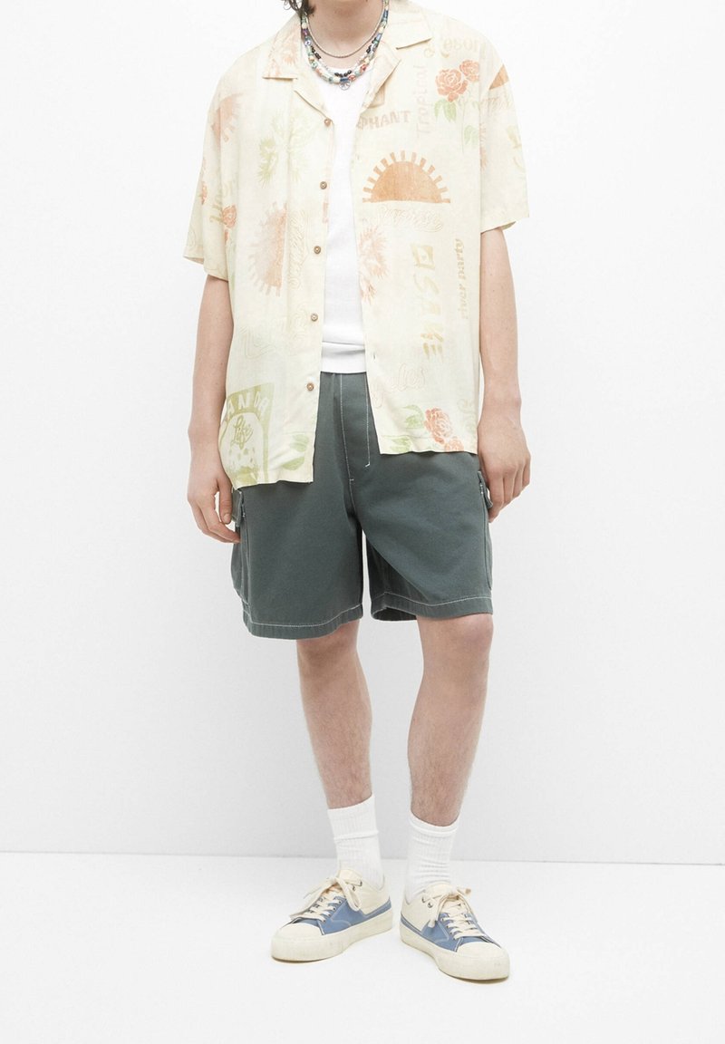 Camicetta beige a maniche corte con bottoni e motivo floreale, abbinata a pantaloni corti verdi. Indossata con calzini bianchi e sneakers crema con dettagli blu.