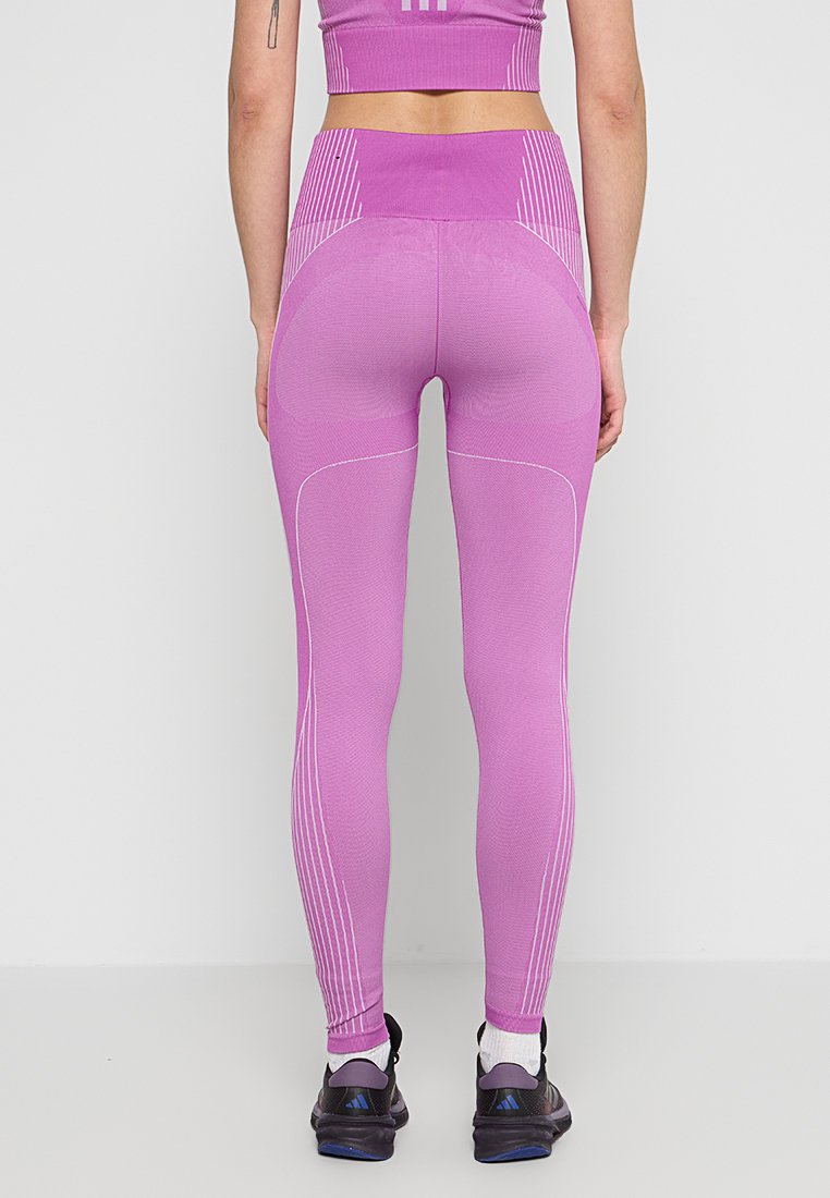 Leggings roses ajustés en tissu texturé, avec des rayures blanches verticales à la taille et sur les côtés, associés à des chaussures de sport noires.