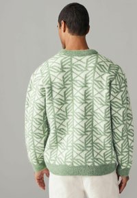 Zöld és fehér mintás pulóver kerek nyakkal és bordás mandzsettával, geometrikus mintázattal az egész felületen. Puha, texturált anyag.