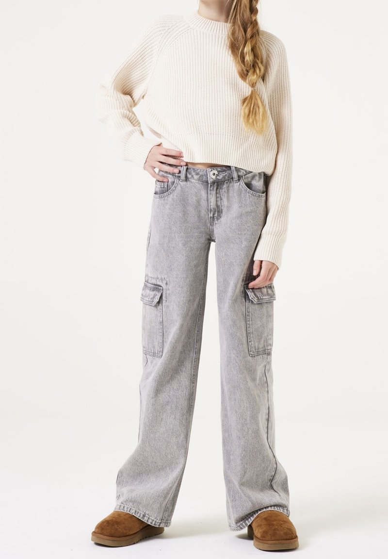 Pull en tricot beige clair, pantalon cargo gris à jambe large avec poches latérales, et pantoufles en peau de mouton brunes. Matériaux unis et texturés.