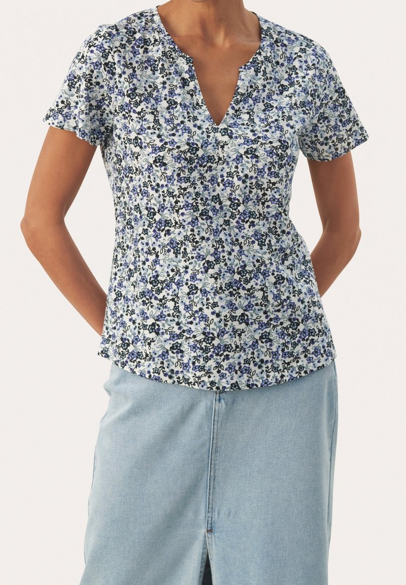 Mujer vistiendo una blusa floral de manga corta con flores azules, negras y blancas, combinada con una falda de mezclilla azul clara con una abertura en la parte frontal.