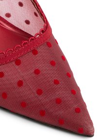 Zapato rojo de punta afilada hecho de malla con un patrón de lunares. Presenta un borde ondulado y paneles transparentes. Textura suave en general.