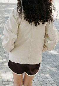 Beige oversized jas met verlaagde schouders, gecombineerd met bruine shorts met witte afwerking, gefotografeerd op een patroon van stoeptegels.