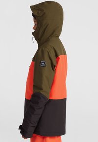 Veste isolante colorée avec capuche vert olive, section supérieure orange et section inférieure noire. Comprend un patch logo et des poignets réglables.