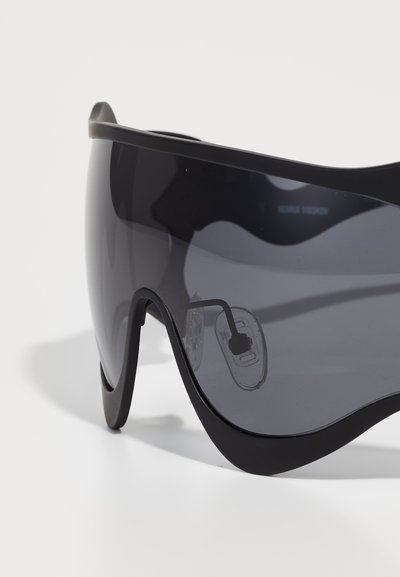 Lunettes de sport noires avec un design enveloppant, verres teintés gris et coussinets de nez transparents. La monture présente une finition lisse et épurée.