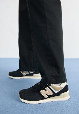 Zwarte sneaker met gestructureerde stof, crème accenten en een prominente grijze "N"-logo. Geplaatst op een blauwe carpet, gecombineerd met zwarte broek.