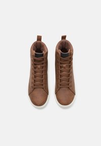Brune høyhælte sneakers laget av teksturert syntetisk lær med flate snører, en hvit gummisåle og en stoffforing.
