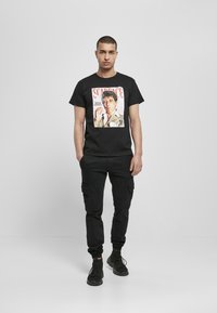 Black t-shirt featuring a prominent graphic print of a character with a light background. Worn with black cargo pants and dark footwear.