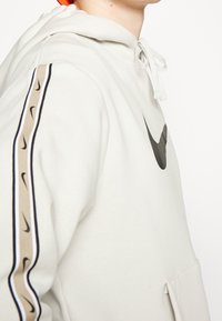 Person klädd i en beige Nike-hoodie med svart swoosh-logotyp på bröstet och en beige rand med mindre svarta swooshes på ärmen.