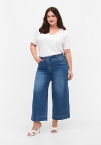 Losse blauwe denim jeans met een hoge taille en zichtbare naden. Gecombineerd met een witte V-hals t-shirt en witte sandalen met hakken.