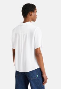 Donna che indossa una blusa bianca corta e larga e jeans blu, vista da dietro su uno sfondo chiaro e semplice.