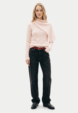 Frau trägt einen hellrosa flauschigen Pullover mit asymmetrischem Ausschnitt, schwarze Jeans mit rotem Gürtel und schwarze Loafers, stehend vor weißem Hintergrund.