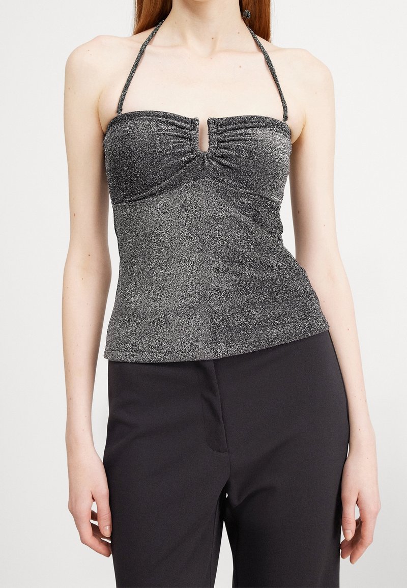 Kobieta w świecącej srebrnej bluzce na ramiączkach z marszczeniem na środku i czarnych spodniach, pokazana od ramion do bioder na białym tle.