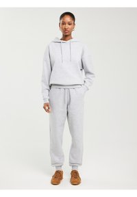 Jauna moteris stovi dėvėdama šviesiai pilką džemperį su gobtuvu ir sportines kelnes bei rudus sportinius batelius ant balto fono.