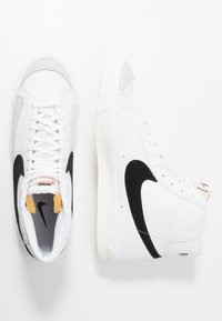 White high-top sneakers with a black Nike swoosh, suede grey accents, white laces, textured sole, and padded tongue.