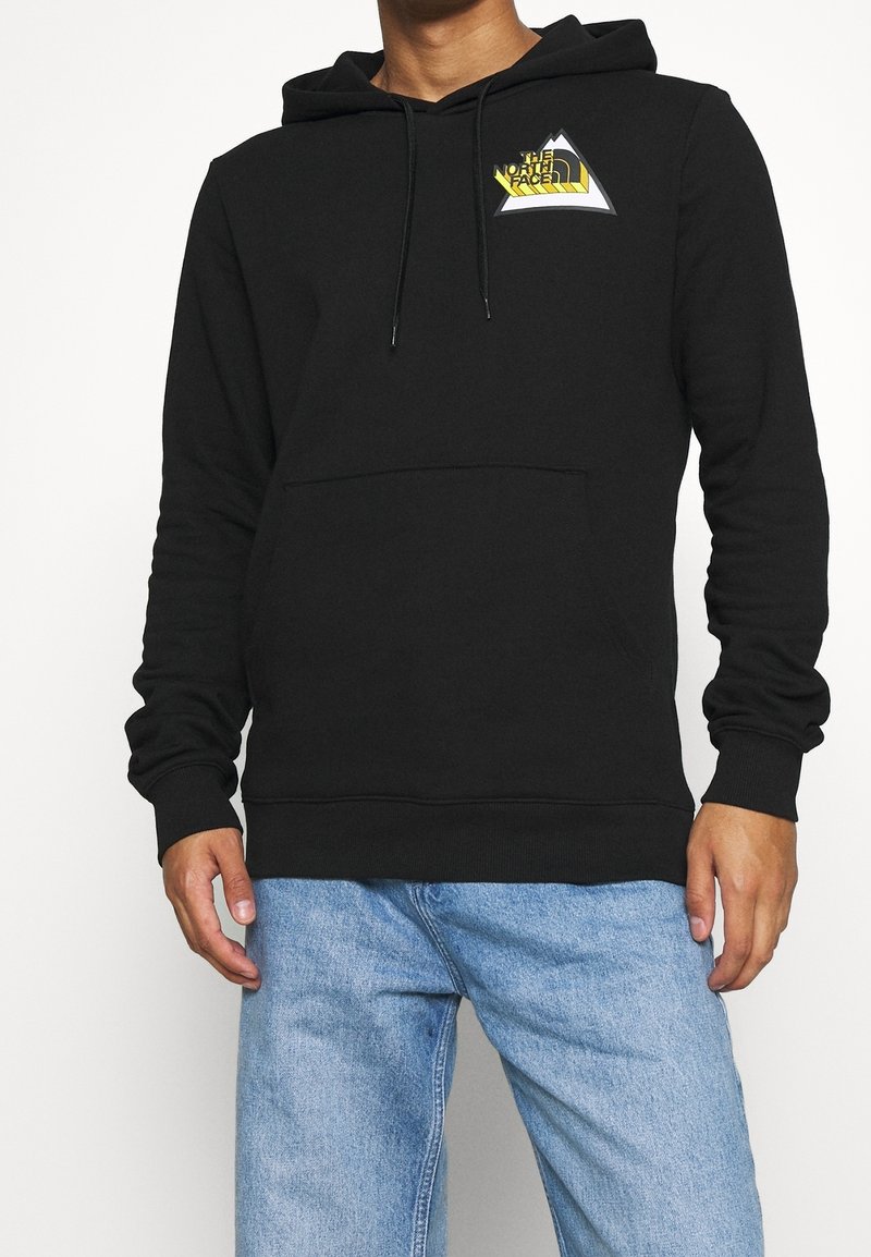 Homme portant un sweat à capuche noir avec un logo triangulaire jaune et blanc sur la poitrine, associé à un jean en denim bleu clair, sur fond blanc.