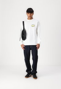 Hvid langærmet skjorte med trykt logo, parret med mørkeblå jeans og sorte sneakers. En lille sort crossbody taske hænger fra skulderen.