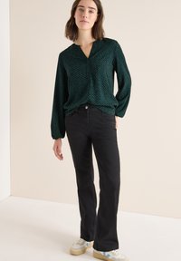 Dunkelgrün gemusterte Bluse mit V-Ausschnitt und langen Ärmeln, kombiniert mit schwarzen Schlagjeans und hellen Sneakers.