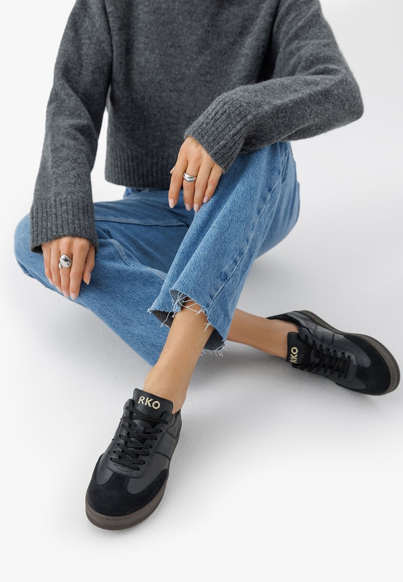 Person som sitter på golvet iförd blå jeans med fransar, mörkgrå tröja, svarta sneakers med "RKO"-logotyp och silverringar på båda händerna.