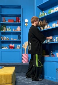 Svart overdimensjonert skjorte og svarte bukser med grønne striper, står foran blå hyller som viser ulike skjønnhetsprodukter og dekorasjoner.