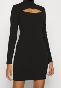 Femme portant une robe noire ajustée à manches longues avec un décolleté en forme de découpe mettant en valeur un petit anneau en métal.