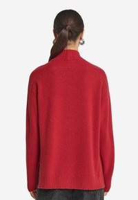 Roter Pullover mit hohem Kragen, gerippten Ärmelbündchen und Saum. Der Stoff wirkt weich mit einer strukturierten Oberfläche. Hat eine lässige Passform und seitliche Schlitze.