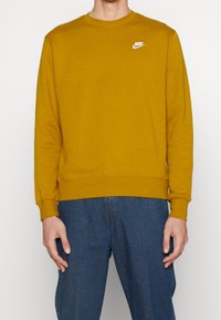 Gul Nike sweatshirt med långa ärmar, rundad hals, ribbade muddar och nederkant, med en vit logotyp på bröstet. Bärs med blå denimbyxor.