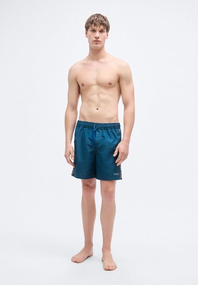 Τιρκουάζ μαγιό σορτς με κορδόνι στη μέση, πλευρικές τσέπες και λογότυπο. Το ύφασμα φαίνεται λείο με ελαφρύ υφή.