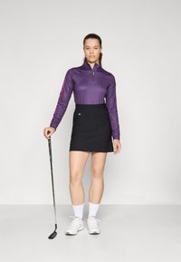 Vijolična dolga majica na zadrgo s dolgimi rokavi v kombinaciji s črnim golf krilom. Majica ima roza črte na rokavih, model pa drži golf palico.