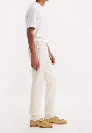 Mężczyzna w białej koszulce, kremowych jeansach z prostymi nogawkami, brązowych mokasynach z zamszu oraz z widocznymi tatuażami na ramionach, stoi bokiem na tle białego tła.