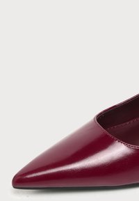 Gros plan d'une chaussure en cuir rouge foncé brillante à bout pointu avec des coutures visibles sur un fond clair.