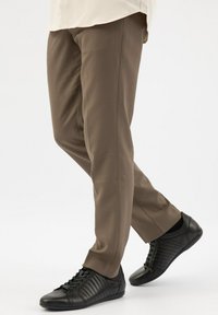 Pantaloni marroni su misura realizzati in tessuto liscio, abbinati a scarpe da ginnastica nere a bassa punta con sottili dettagli a coste. Sfondo bianco uniforme.