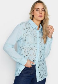 Camicia azzurra chiara con design a bottoni, caratterizzata da intricate lavorazioni a taglio, maniche lunghe e colletto appuntito, realizzata in tessuto traspirante.