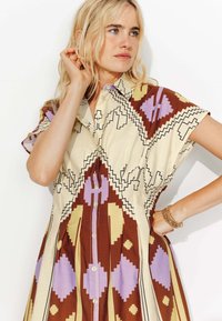 Robe col camisole avec un motif géométrique en marron, crème, violet et jaune. Manches courtes et fermeture à boutons avec jupe plissée.