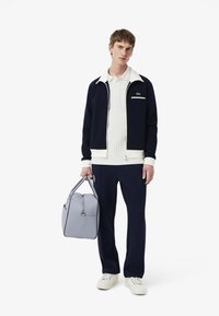 Marinblå zip-up jacka med krämfärgad krage, vit ribbad polo, marinblå byxor och ljusgrå handväska. Avslappnad stil med subtila logotypdetaljer.