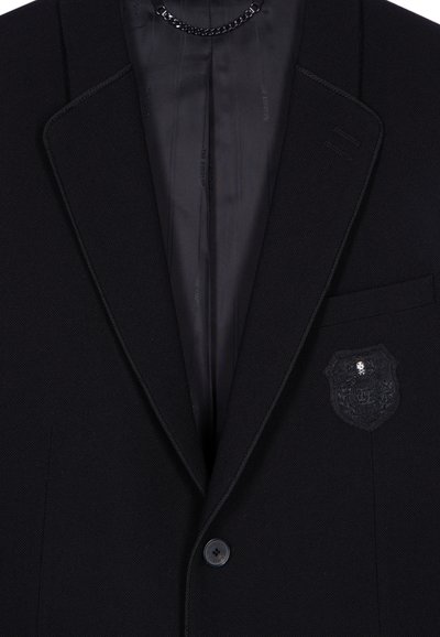 Schwarzer maßgeschneiderter Blazer mit strukturierter Außenseite, satinierter Innenfutter, einem dekorativen Wappen-Patch auf der Brusttasche und einem einzelnen Knopfverschluss.