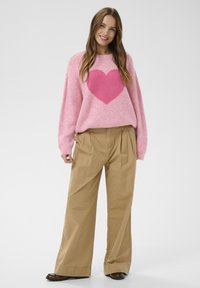 Femme souriante portant un pull rose avec un grand cœur rose foncé, un pantalon large beige et des chaussures marron, debout sur un fond blanc.