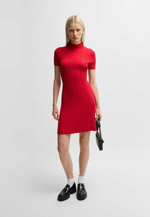 Rotes geripptes Kleid mit hohem Kragen, kurzen Ärmeln und ausgestelltem Saum. Kombiniert mit schwarzen Slippern und einer kleinen schwarzen Handtasche.