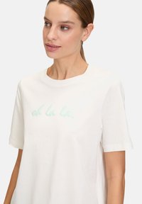 Hvid bomulds-T-shirt med rund hals, korte ærmer og en lysegrøn broderet tekst, der siger "oh la la."