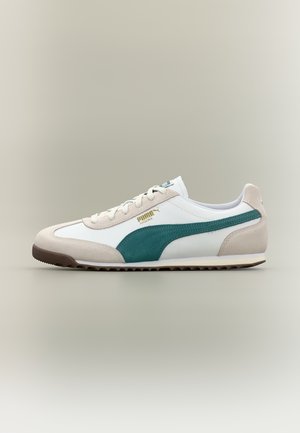 Bijela Puma Arizona tenisica s bež brušenim detaljima, tirkiznom bočnom prugom i smeđim đonom, prikazana u profilu na neutralnoj pozadini.