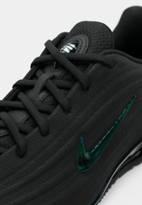 Close-up de sapatilha preta com parte superior texturizada, atacadores pretos e um logo Nike em verde brilhante na lateral.