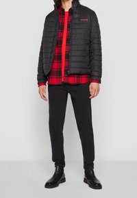 Czarna kurtka puchowa z zamkiem z przodu, z czerwonym logo "HUGO". Pod spodem czerwona i czarna koszula w kratę. Czarne spodnie i błyszczące czarne buty.