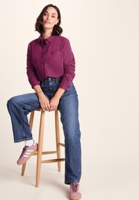 Purple button-up shirt with long sleeves and a chest pocket, paired with high-waisted blue jeans and pink sneakers. Wooden stool backdrop.