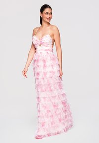 Femme portant une robe à volants à étages rose et blanche à motifs floraux, avec de fines bretelles, souriante et regardant sur le côté sur un fond clair uni.