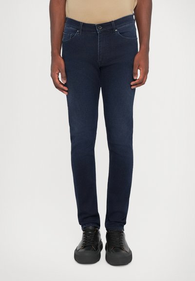 Jeans de mezclilla azul oscuro con corte ajustado, que presentan bolsillos delanteros, trabillas para cinturón y detalles de costura mínimos. Combinados con zapatillas negras.