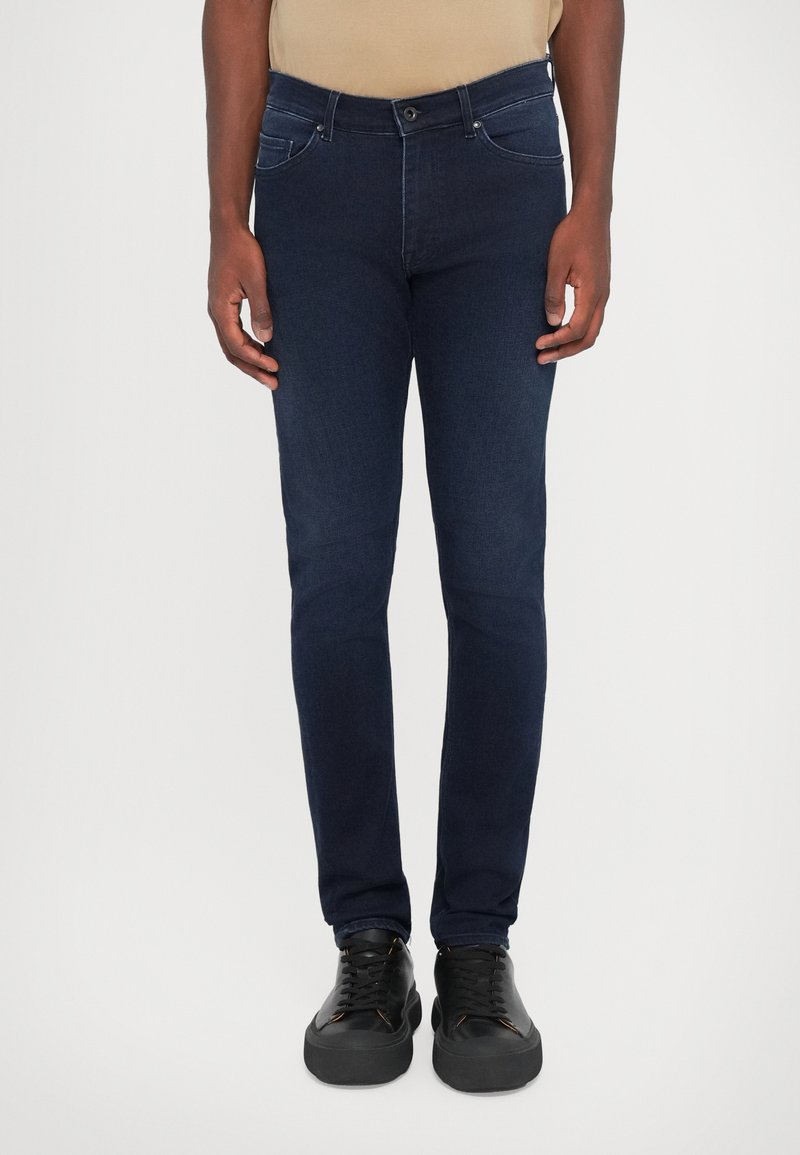 Jeans din denim albastru închis, cu croială slim, având buzunare frontale, curea și detalii minime de cusătură. Asortați cu adidași negri.