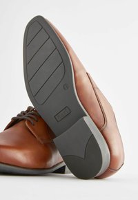Chaussure en cuir marron avec une surface lisse et un bout arrondi ; design à lacets. Semelle en caoutchouc noire avec des rainures horizontales et un détail de marque.