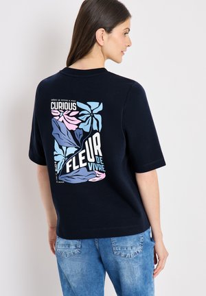Donna con lunghi capelli scuri che indossa una t-shirt blu navy con stampa floreale pastello e testo francese "Curious Fleur de Vivre" sul retro, abbinata a jeans blu.